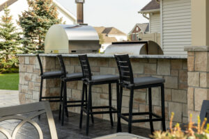A beautifully designed outdoor covered patio by R.E. McNamara Inc., featuring a stainless steel grill, stylish bar seating, a modern brick oven, and a cozy fire pit with comfortable seating area. The atmosphere is warm and inviting, perfect for residential gatherings and entertaining guests.