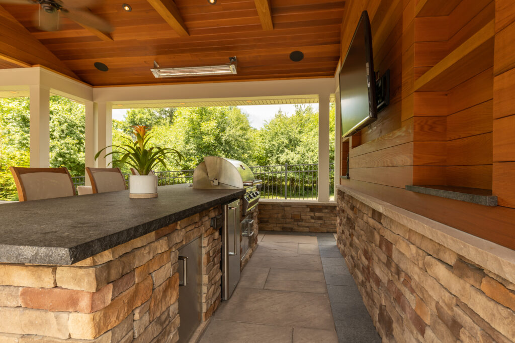 A covered outdoor patio featuring a cooking station with a grill, built-in minifridge, four bar stools, and a mounted TV