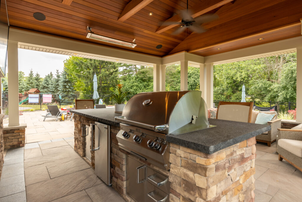 A covered cooking area with a grill, built-in minifridge, and seating for four by R.E. McNamara Inc.