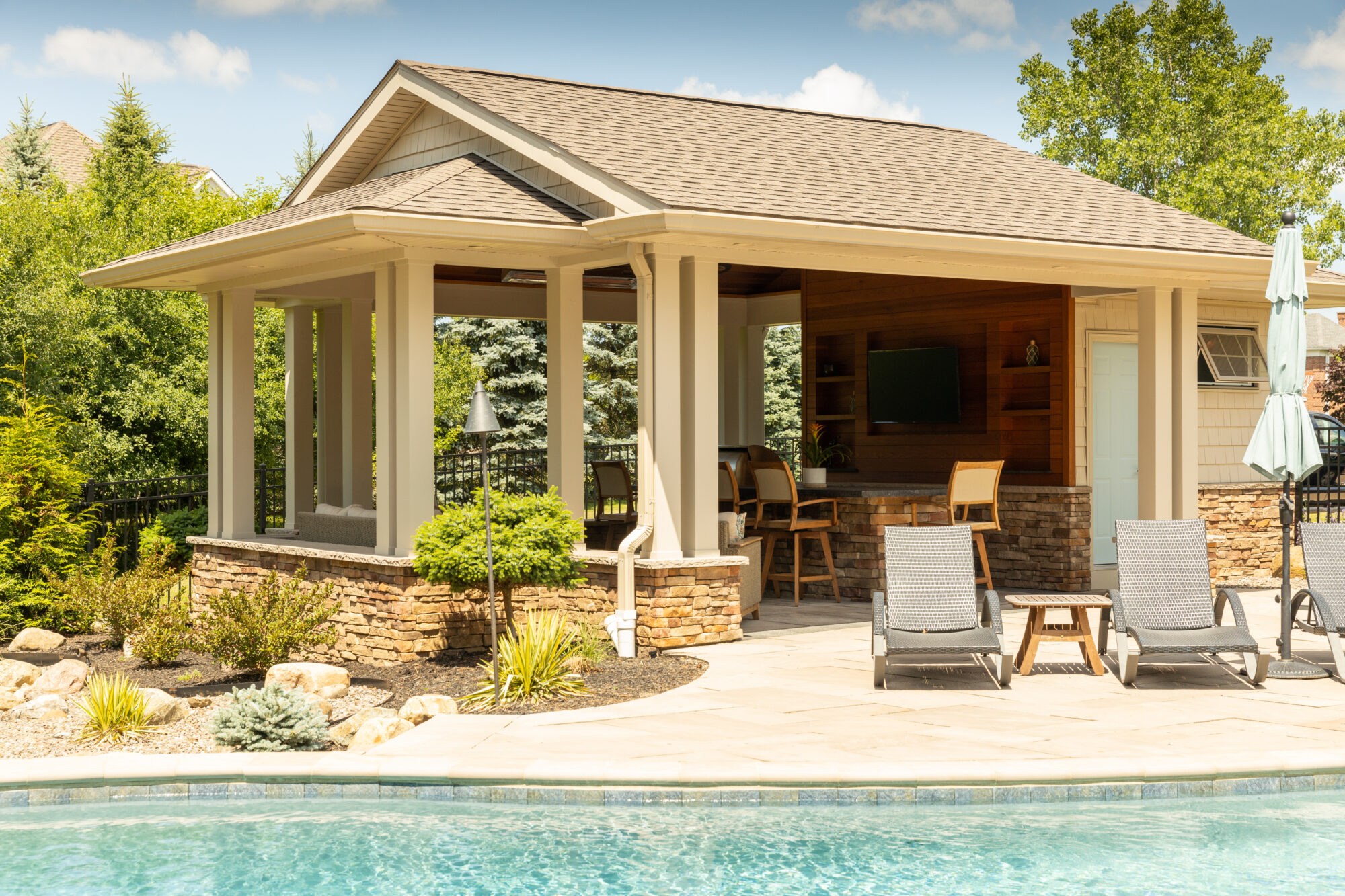 Covered patio space by an inground pool featuring a fully-equipped cooking station, comfortable bar seating, and stylish stone walls enhanced with rich wooden accents