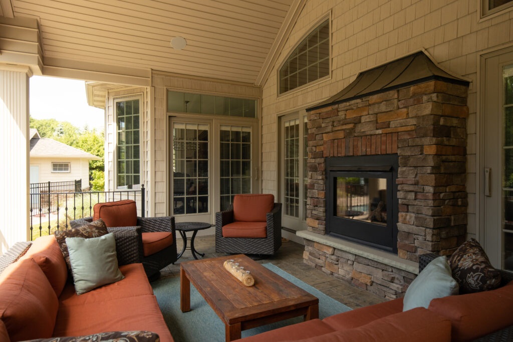 A cozy, covered outdoor patio designed by R.E. McNamara Inc., featuring a stone-covered fireplace and warm-colored patio furniture