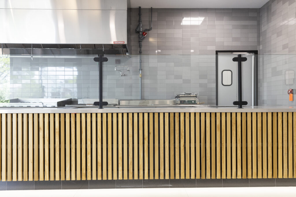 Service counter at Rachel's Mediterranean Grill, featuring a clean food prep area, light wooden accents, bright flooring, and a modern grey tile wall by R.E. McNamara Inc.