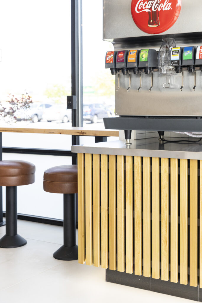 A self-serve soda machine and stool seating area at Rachel's Mediterranean Grill designed by R.E. McNamara Inc., showcasing light wooden accents and bright surfaces.