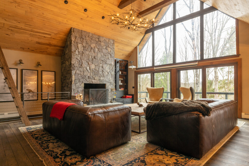 A stunning living room renovation showcasing a centerpiece stone fireplace, contrasting with the dark hardwood floors and warm wood-paneled ceiling, all bathed in sunlight from large surrounding windows.
