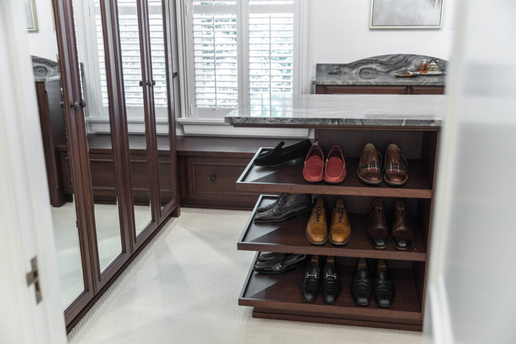 A luxurious residential closet renovation, highlighting white walls and deep brown wooden shelves, accompanied by a functional shoe-storage island in the center.