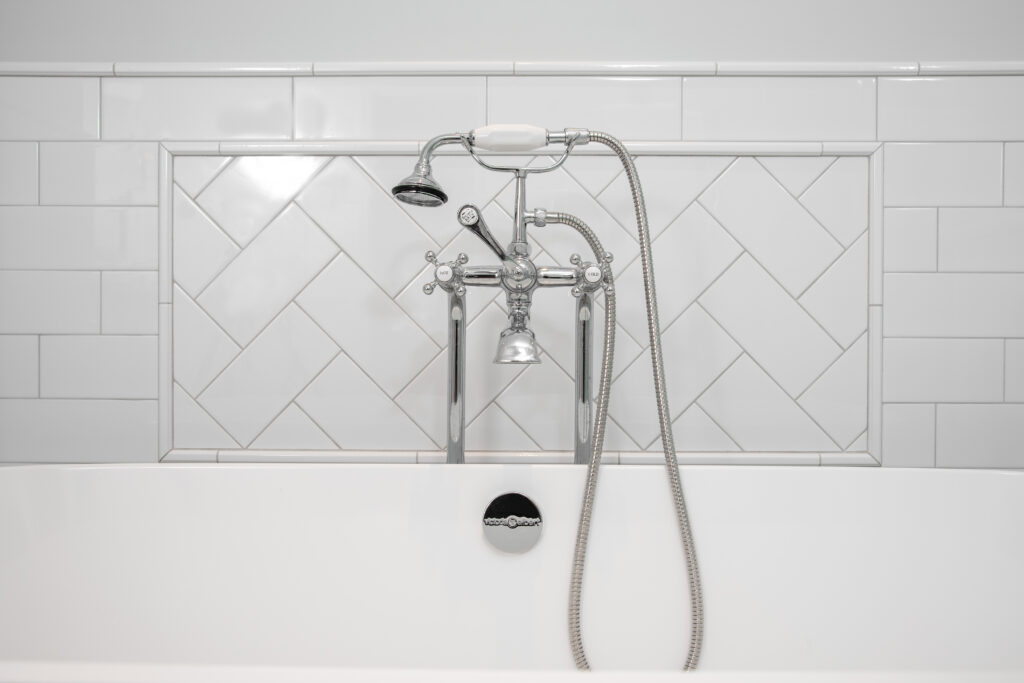 Close-up of sleek bathtub fixtures, including handle and head with handheld attachment, complemented by a stylish tiled backsplash above the tub area