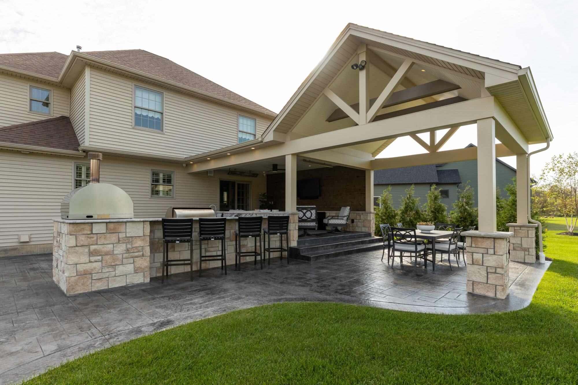 An inviting and beautifully designed outdoor covered patio area featuring a stainless steel grill, stylish bar seating with comfortable cushions, a modern built-in oven, and a cozy round fire pit with surrounding chairs, showcasing R.E. McNamara Inc.'s expertise in residential renovation.