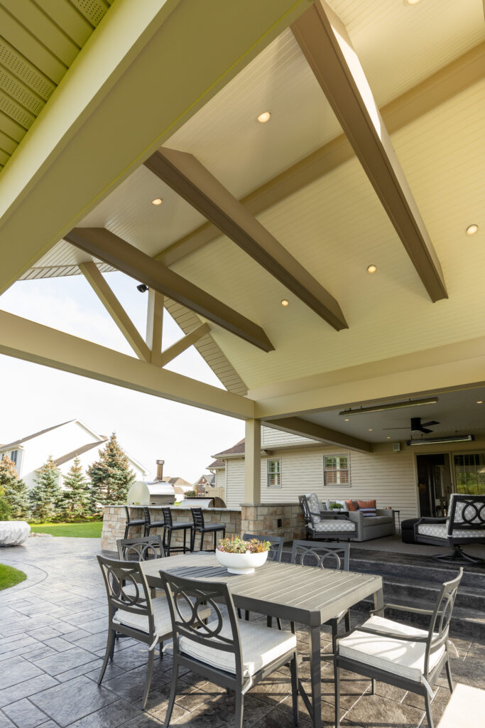 An inviting and beautifully designed outdoor covered patio area featuring a stainless steel grill, stylish bar seating with comfortable cushions, a modern built-in oven, and a cozy round fire pit with surrounding chairs, showcasing R.E. McNamara Inc.'s expertise in residential renovation.